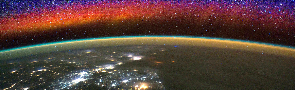 a red band of glowing air is seen above a thin green band above a nighttime scene on Earth