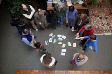 overhead photo of a group of people standing in a circle around cards on floor